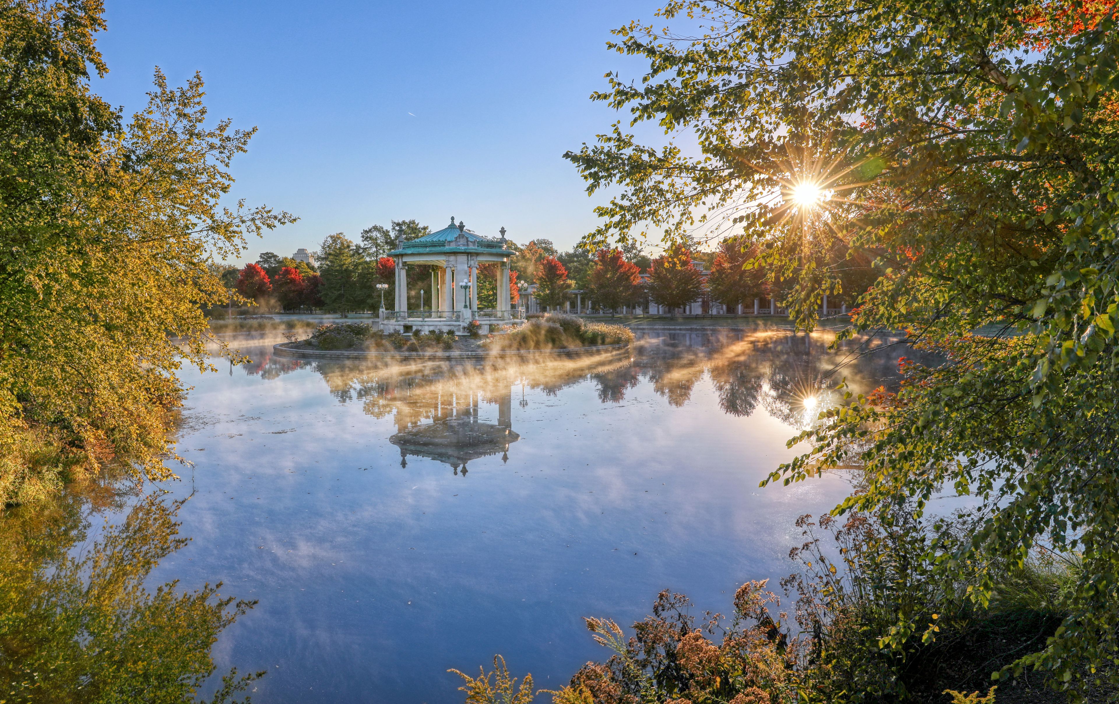 A gazebo by a calm lake with autumn foliage and a clear sky.