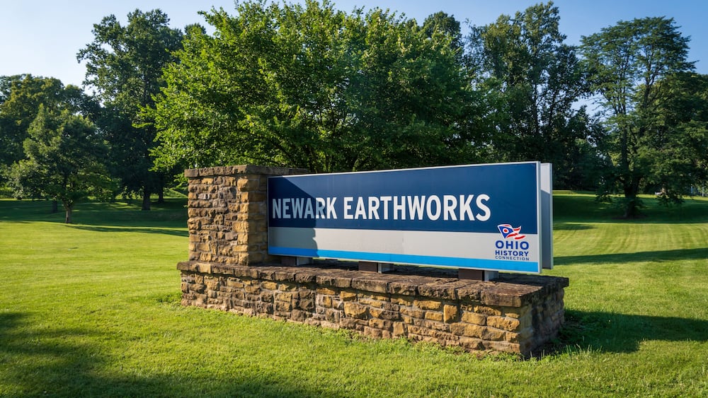 A stone sign with 'Newark Earthworks' and 'Ohio History Connection' text, surrounded by a grassy field and trees.