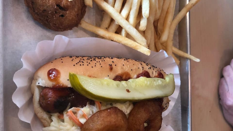 A hot dog with a bun, pickles, coleslaw, cheese, and onions, along with a side of French fries and onion rings.
