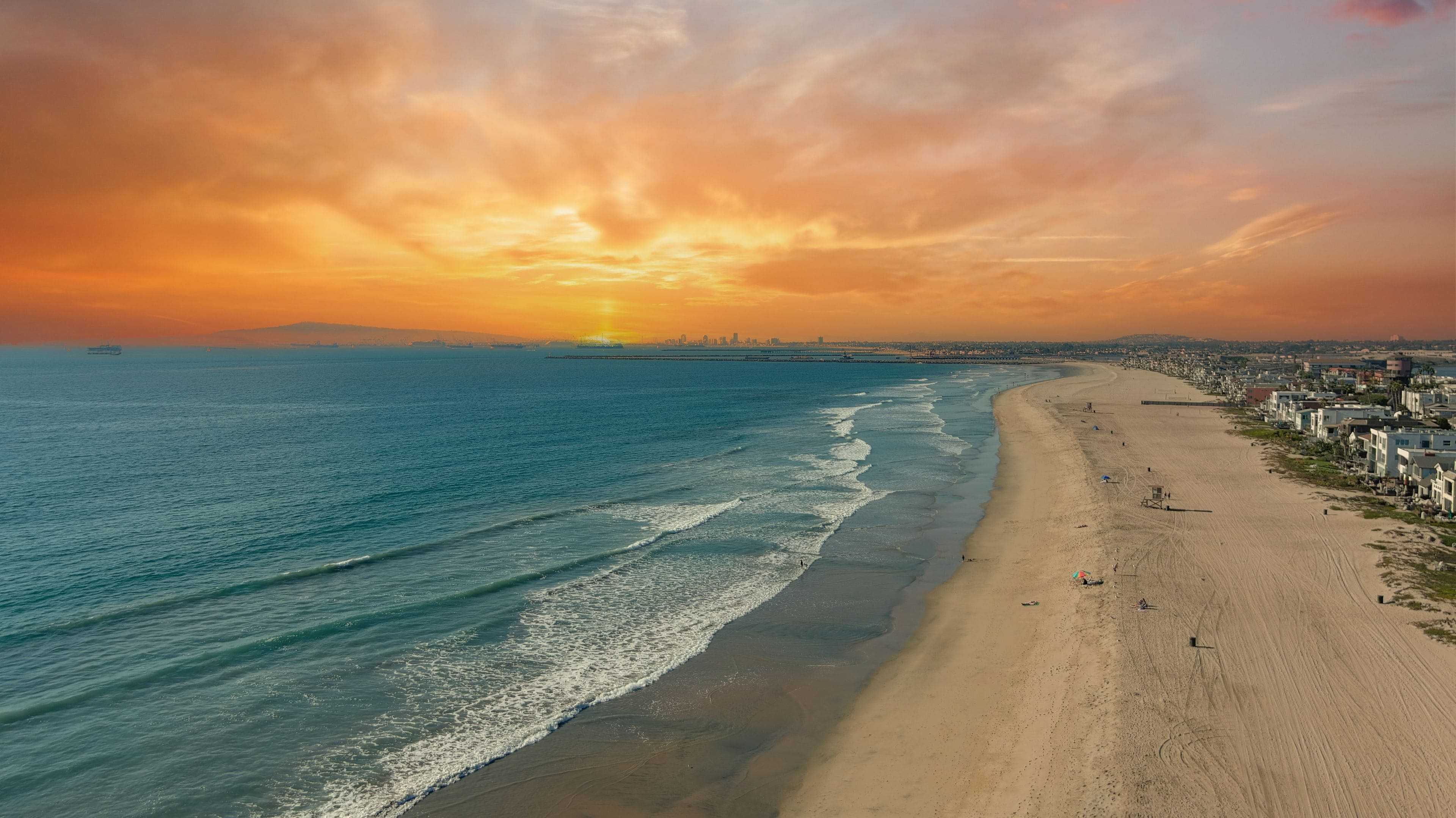 A beach with waves, a sunset, and a city skyline in the distance.