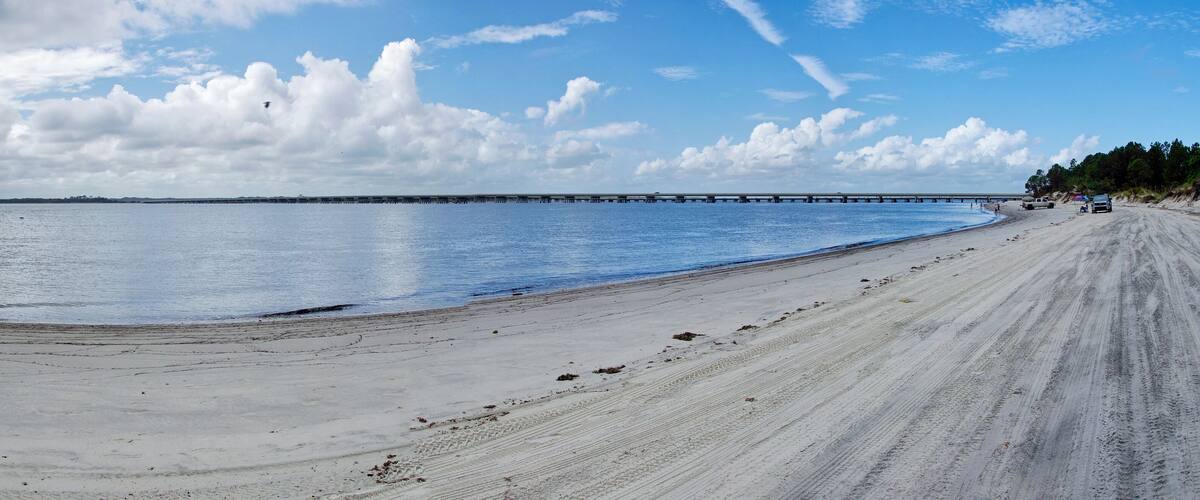A sandy beach with a calm sea, a pier extending into the water, and a clear sky with clouds.