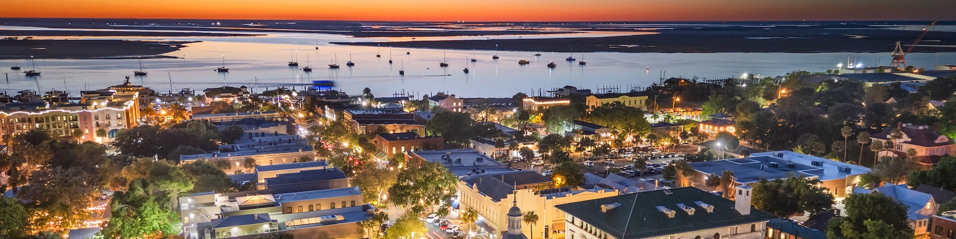A coastal town at dusk with illuminated buildings, a marina with boats, and a prominent clock tower.