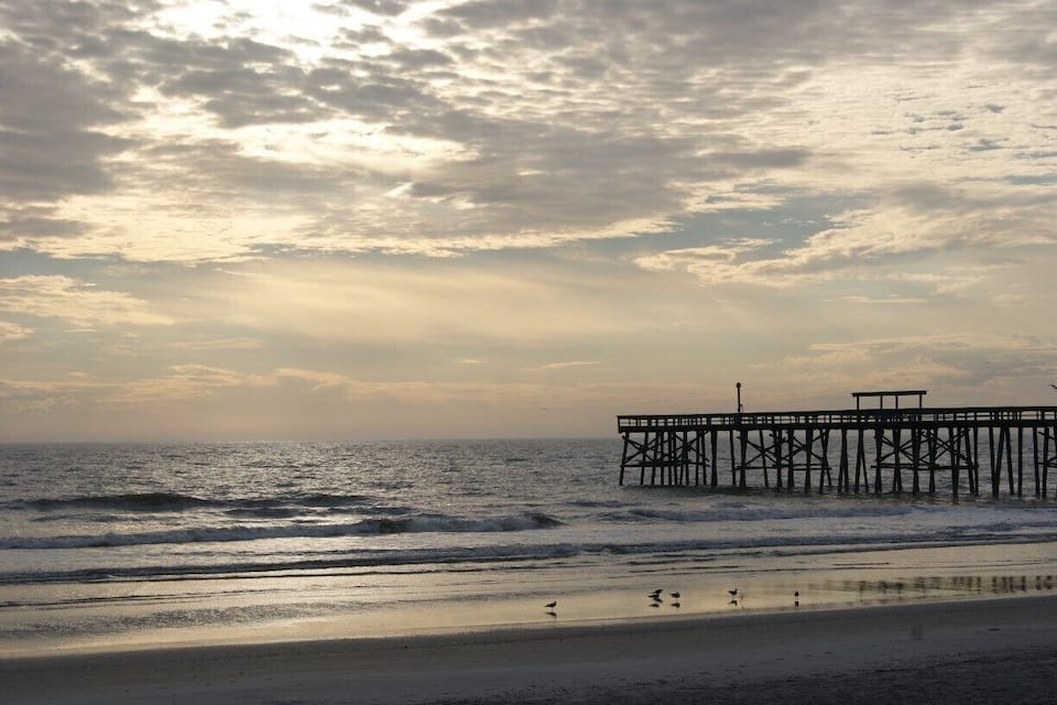 A beach with waves, a pier, and a cloudy sky.