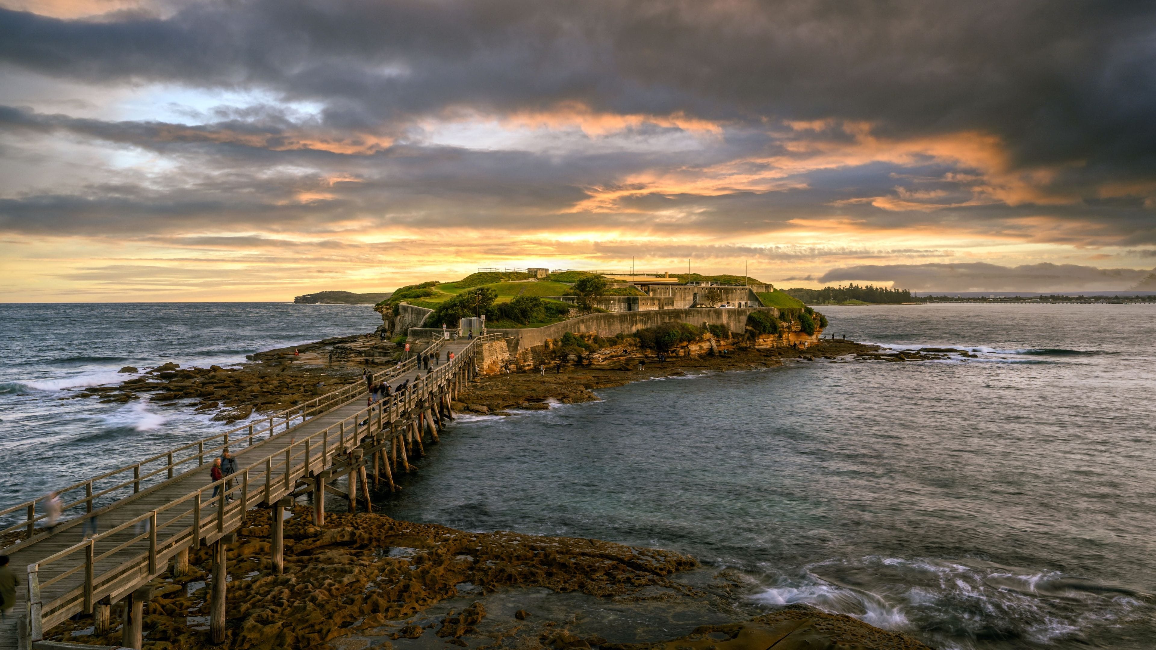 A coastal fortification with a wooden pier extending into the sea, surrounded by rocky terrain and a dramatic sunset sky.