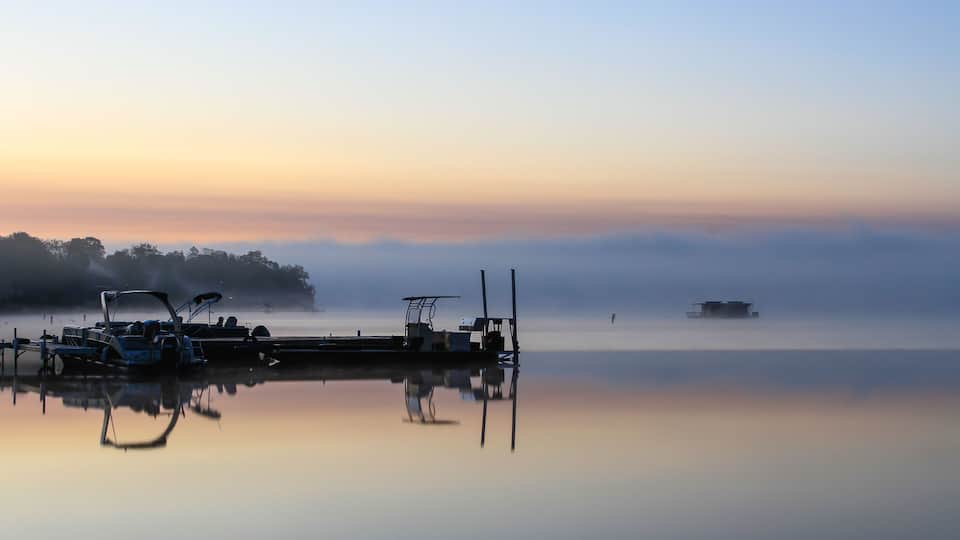 A calm lake at sunrise with boats docked at a pier, surrounded by mist and trees.