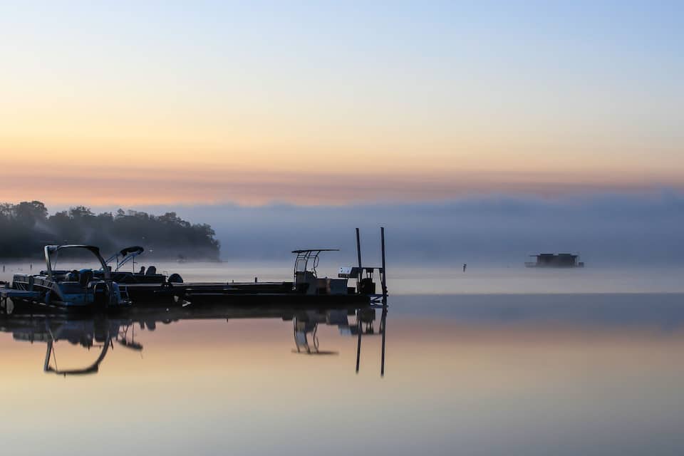 A calm lake at sunrise with boats docked at a pier, surrounded by mist and trees.