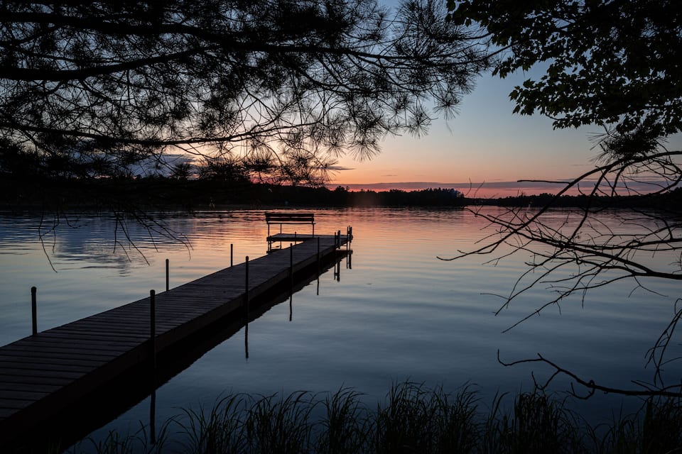 Une jetée en bois s’étendant dans une eau calme au coucher du soleil, entourée d’arbres.