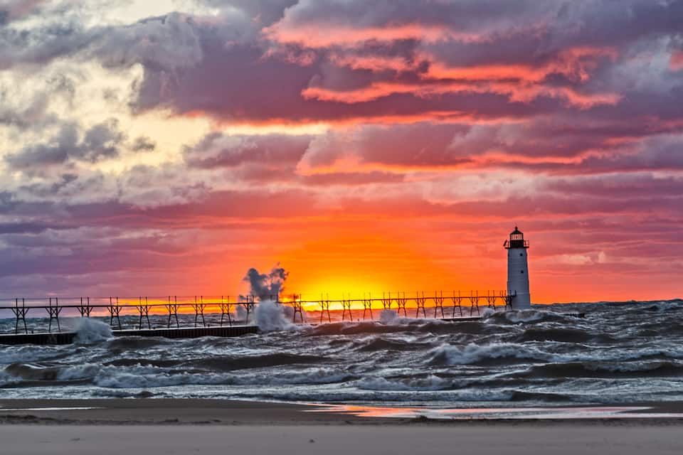 A lighthouse at sunset with waves crashing against a pier.