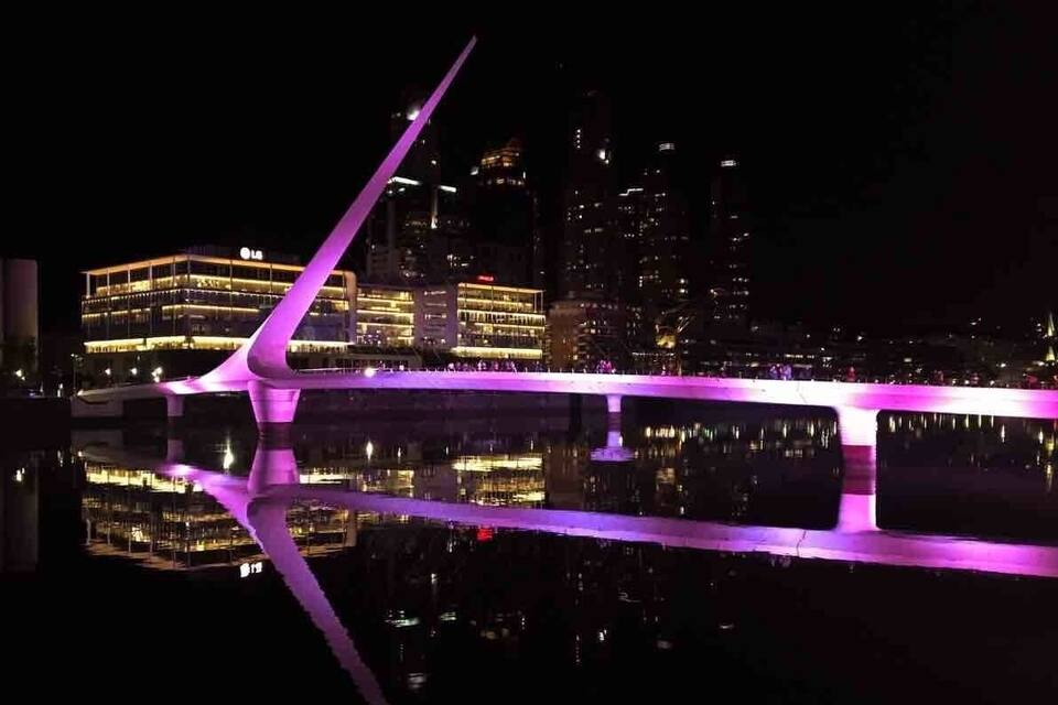 Eine moderne Brücke mit violetter Beleuchtung, eine Stadtlandschaft mit beleuchteten Gebäuden und ein Spiegelbild im Wasser.