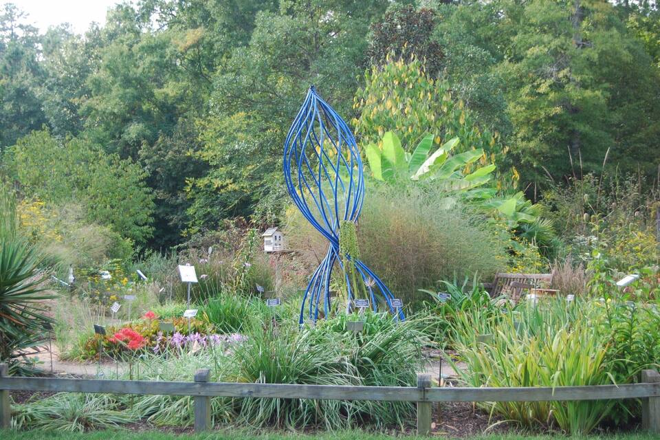 Um jardim com uma escultura de metal azul, diversas plantas e uma cerca de madeira.