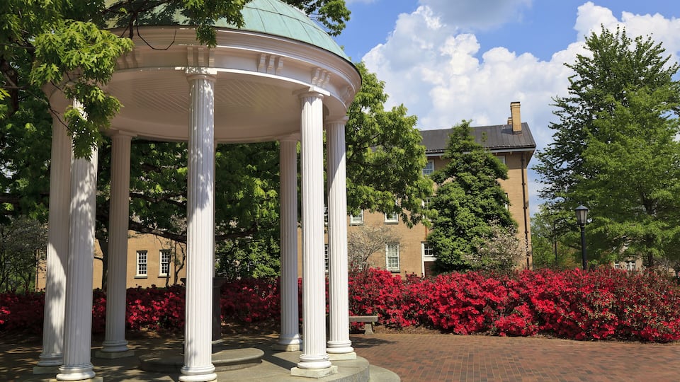 A gazebo with white columns, surrounded by red flowers and trees, in front of a brick building.