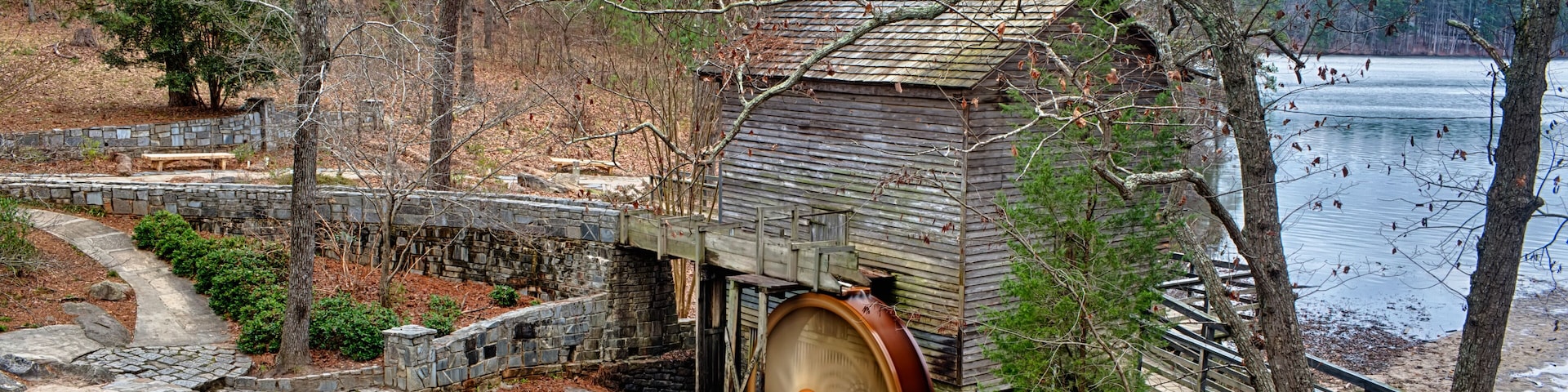 A wooden mill with a large water wheel, a stone bridge, and a lake in the background.