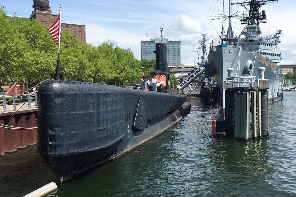 A large naval ship docked at a pier with a cityscape in the background.