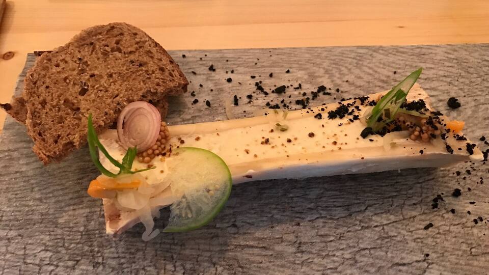 A charcuterie board with a slice of bread, a piece of cheese, cucumber, onion, and garnishes on a slate plate.