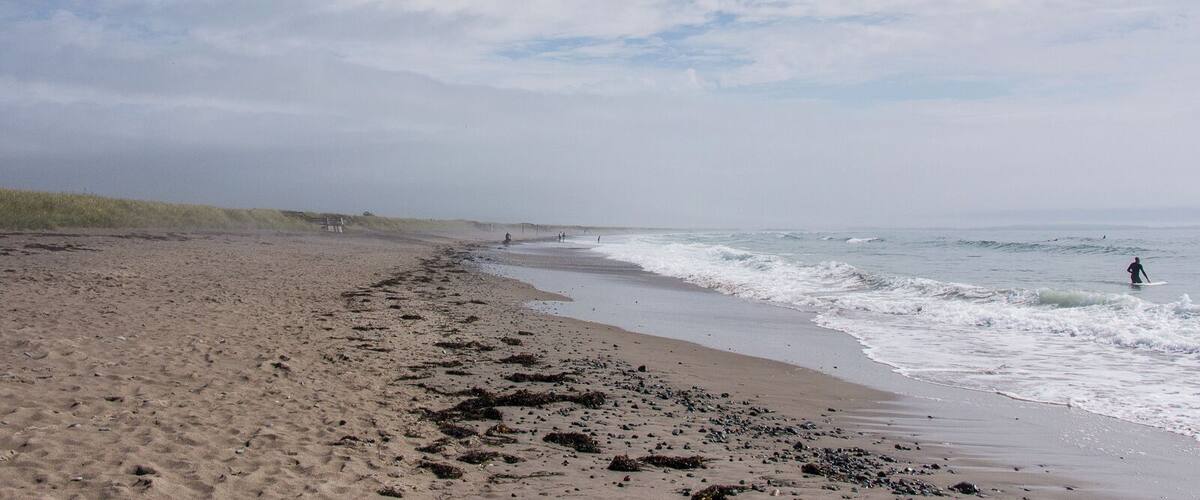 A sandy beach with waves, a clear sky, and a distant person surfing.