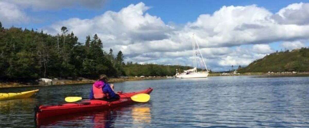 A person kayaking on calm water with a sailboat in the background and a forested shoreline.
