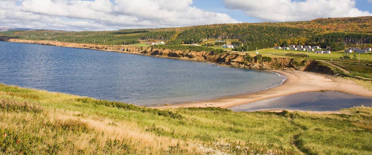 A coastal landscape with a sandy beach, calm water, grassy hills, and scattered houses.