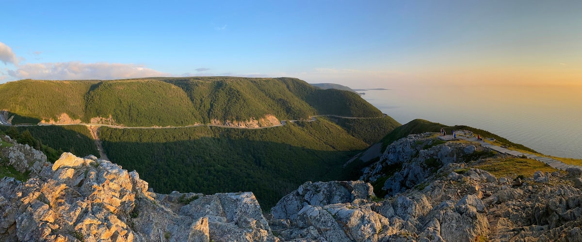 A mountainous landscape with a winding road, a wooden walkway, and a sunset over a body of water.