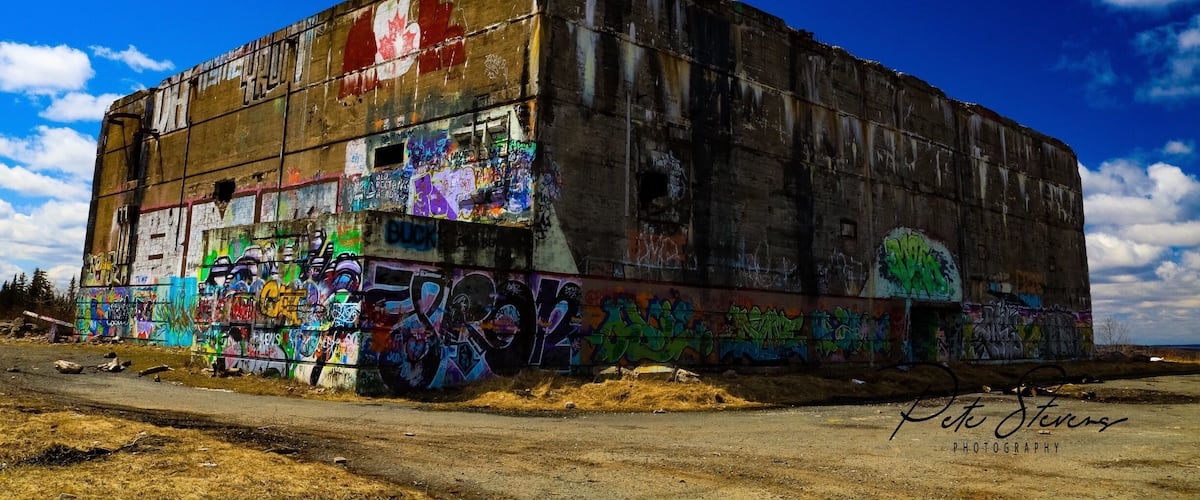A large, graffiti-covered industrial building with a blue sky and clouds in the background.
