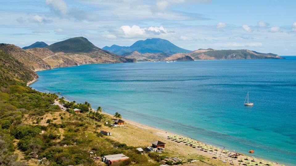 A coastal landscape with a beach, clear blue waters, and distant mountains.