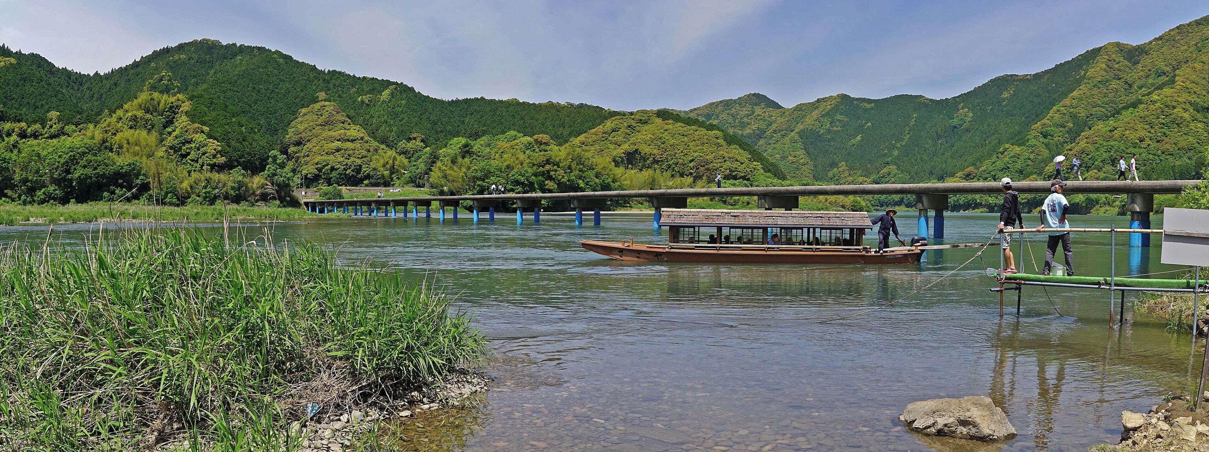 A river with a wooden boat, a bridge, and lush green hills in the background.