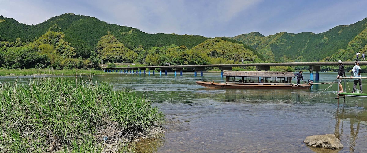 A river with a wooden boat, a bridge, and lush green hills in the background.