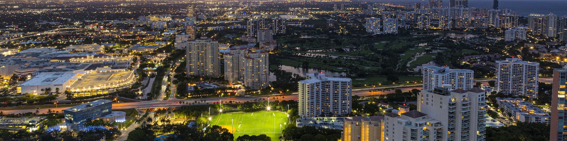 Aerial view of illuminated buildings casting golden glows against the deepening blues of twilight in the vibrant cityscape, Aventura, Florida, United States.
