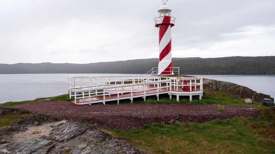 A lighthouse with a red and white striped pattern, a white platform with railings, and a rocky shoreline with a body of water in the background.