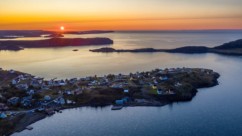 A coastal village at sunset with a lighthouse, calm waters, and distant landforms.