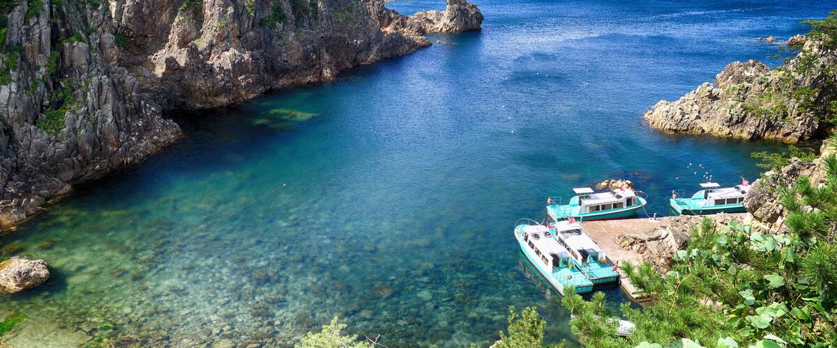 A coastal area with clear blue waters, rocky cliffs, and turquoise boats docked near the shore.
