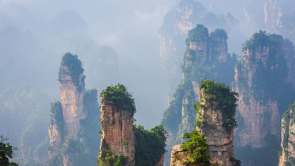 Towering limestone cliffs with dense greenery at their peaks, surrounded by misty mountains.