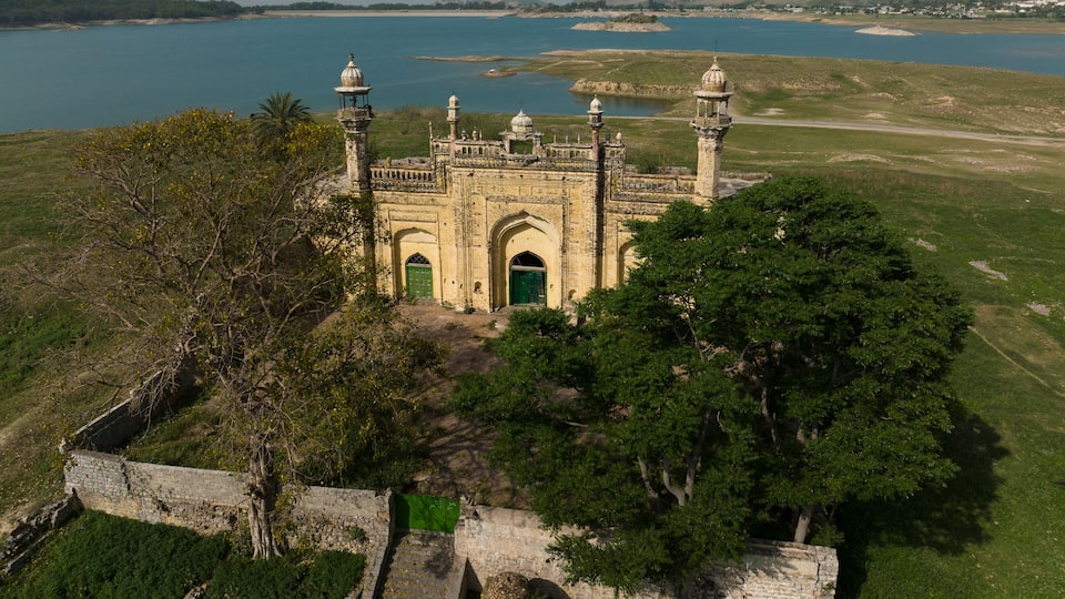 An ancient fortress with multiple towers and a large green door, surrounded by lush greenery and a lake in the background.