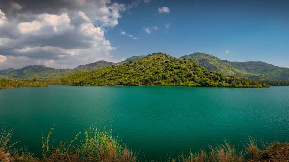A turquoise lake surrounded by green hills and mountains under a partly cloudy sky.