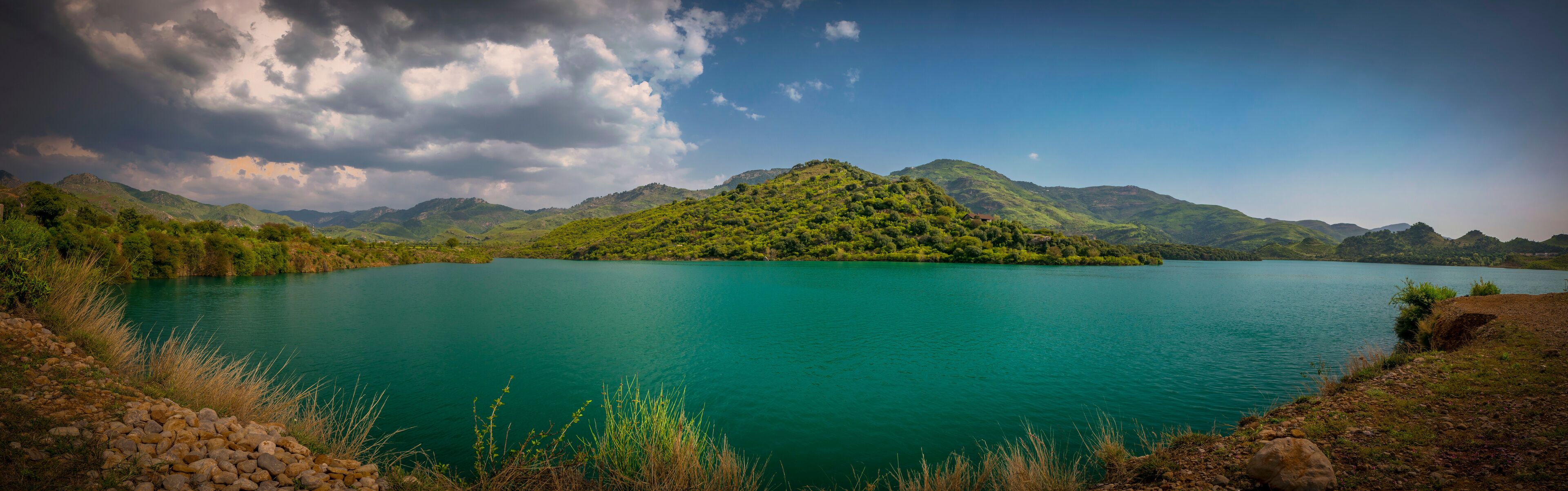 A turquoise lake surrounded by green hills and mountains under a partly cloudy sky.