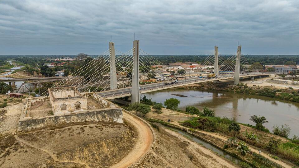 A cable-stayed bridge over a river with a hilly landscape and a small town in the background.
