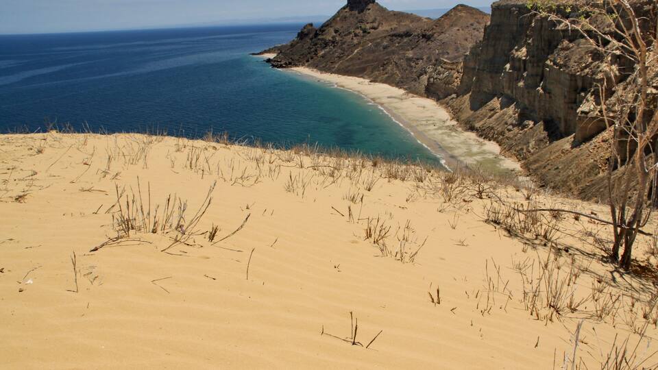 A coastal landscape with a sandy beach, clear blue waters, and rocky cliffs.