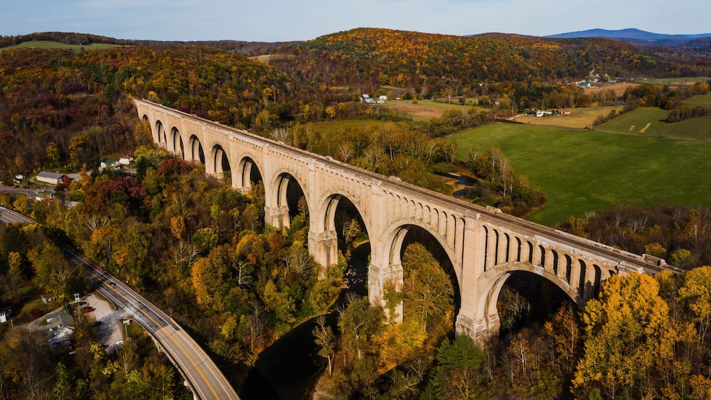 A stone viaduct spanning a river, surrounded by autumn foliage and hills.