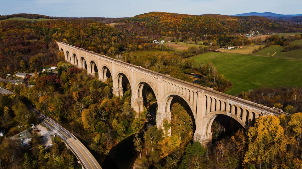A stone viaduct spanning a river, surrounded by autumn foliage and hills.