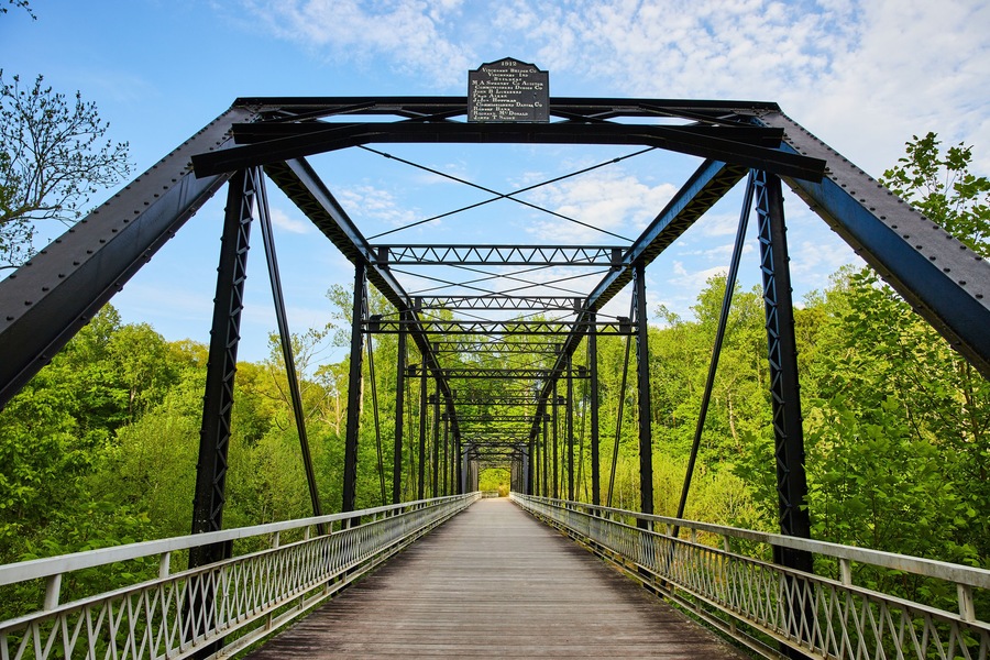 Lush, green forest wall behind large dark metal bridge, black beams, blue sky, woodland background