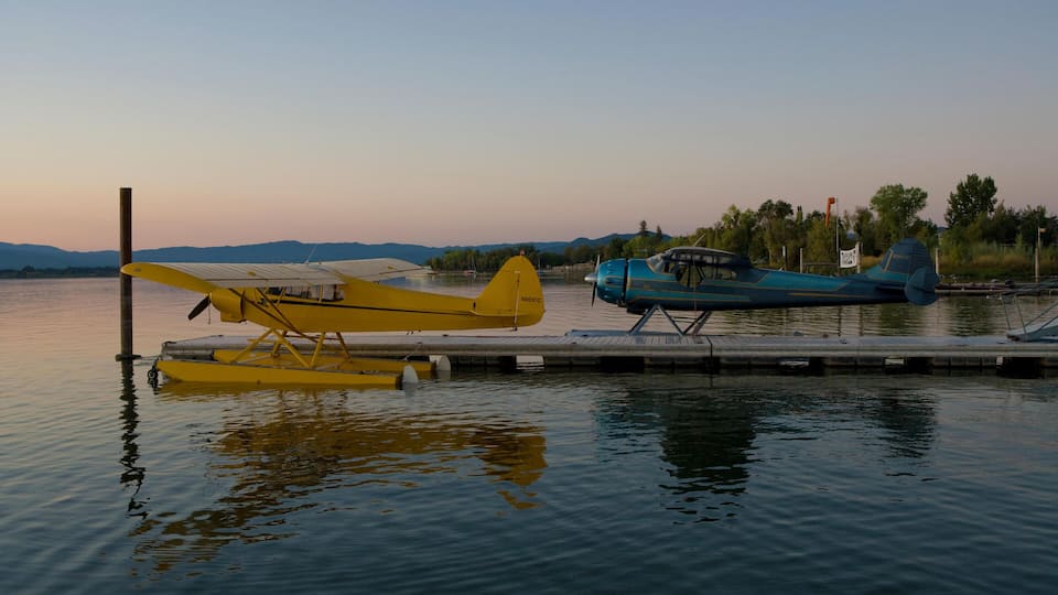 Two seaplanes docked at a pier with calm water and a sunset in the background.