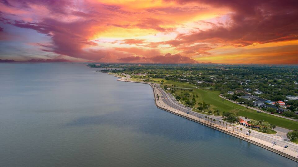 A coastal landscape with a calm sea, a curved road, and a distant city skyline under a colorful sunset sky.