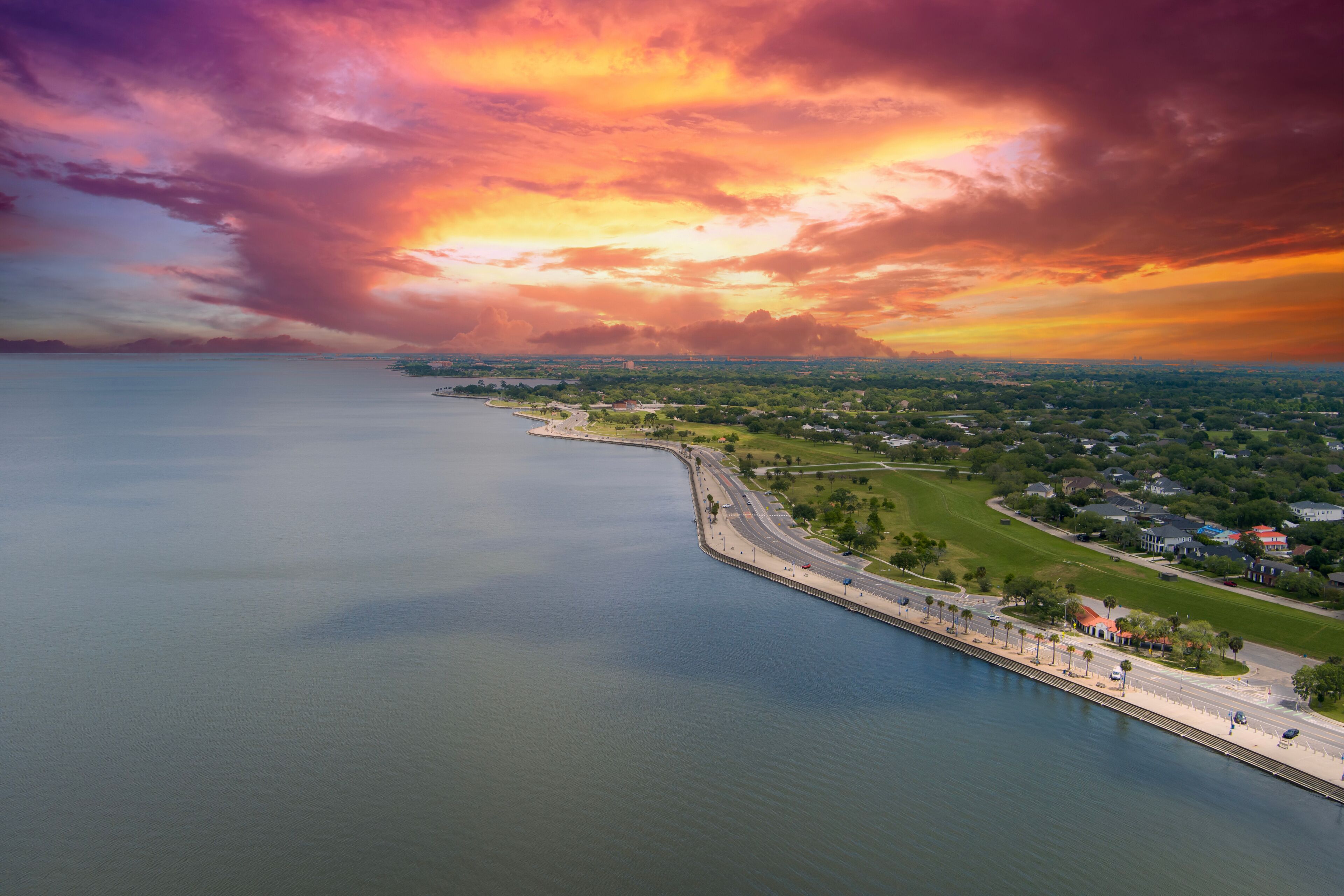 A coastal landscape with a calm sea, a curved road, and a distant city skyline under a colorful sunset sky.