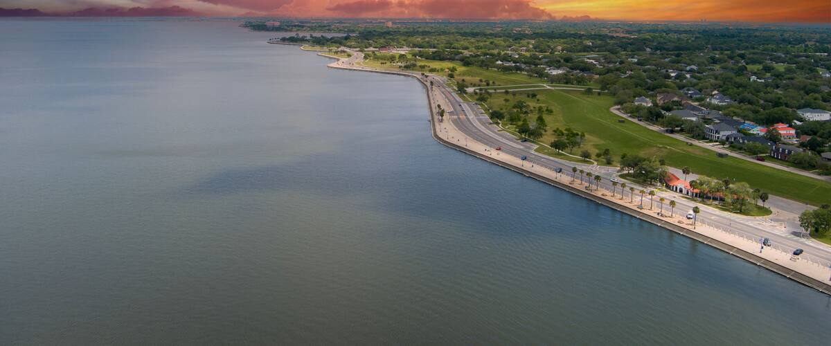 A coastal landscape with a calm sea, a curved road, and a distant city skyline under a colorful sunset sky.
