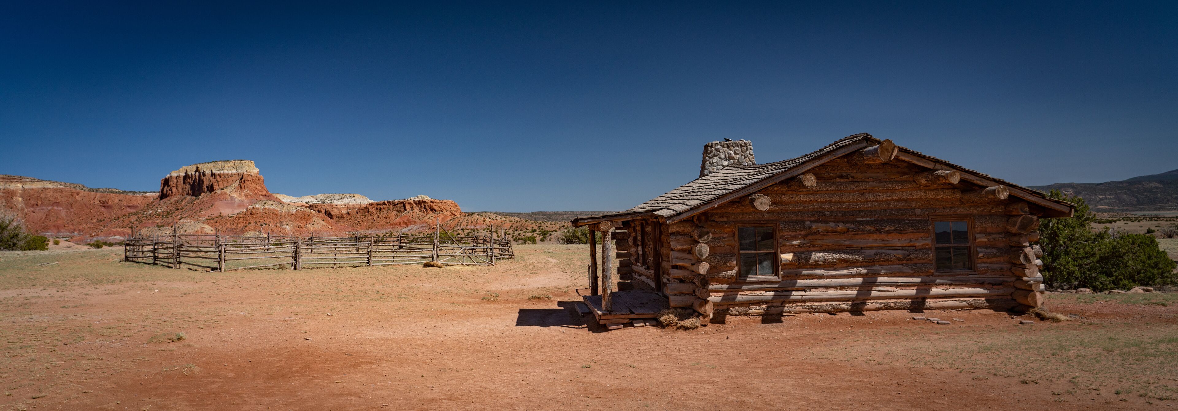A rustic log cabin with a stone chimney, surrounded by a wooden fence and a red rock formation in the background.