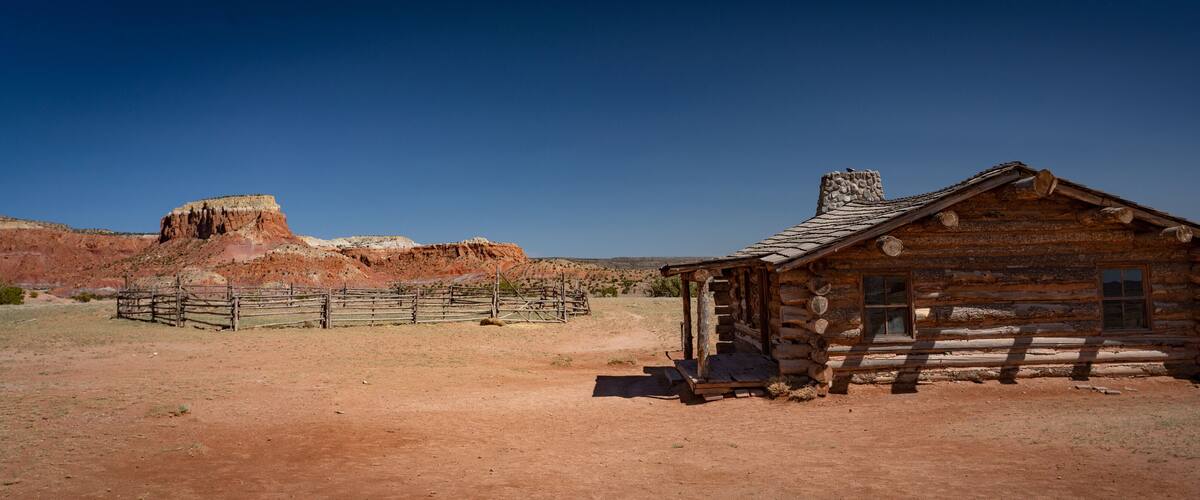 A rustic log cabin with a stone chimney, surrounded by a wooden fence and a red rock formation in the background.