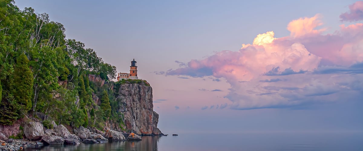 A coastal landscape with a lighthouse on a rocky cliff, calm sea, and a sunset sky.