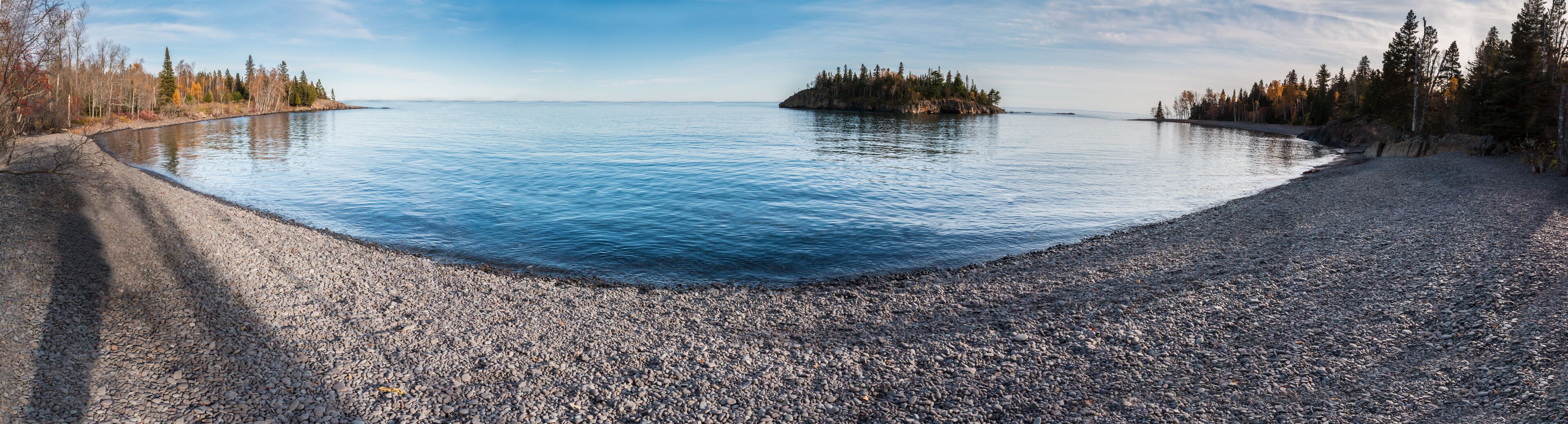 A pebble beach leading to calm waters with a small island and forested shoreline.