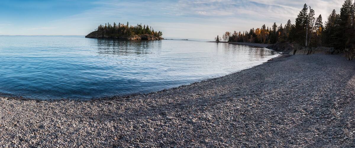 A pebble beach leading to calm waters with a small island and forested shoreline.