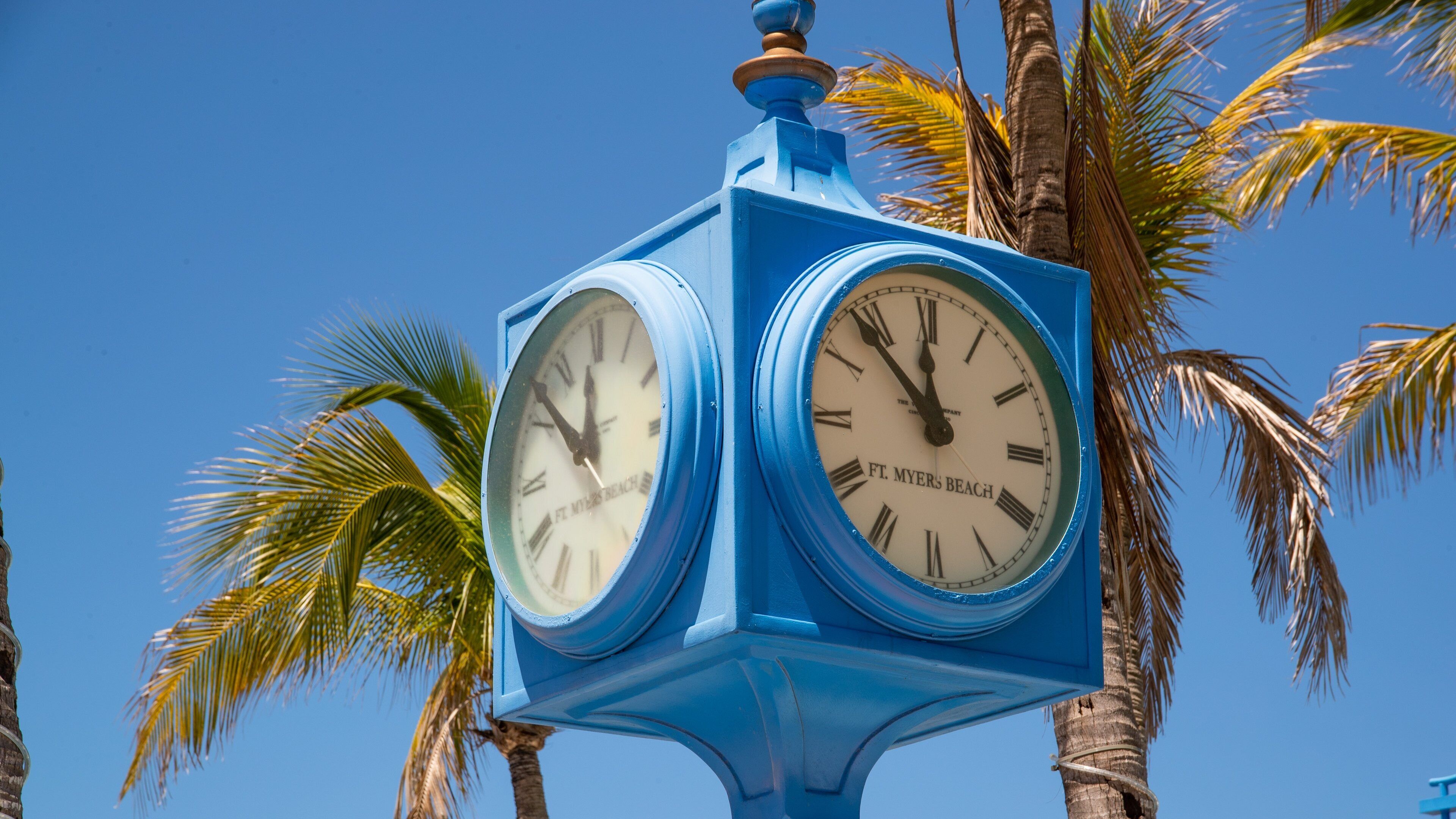 A blue clock with Roman numerals, labeled "Ft. Myers Beach," set against a clear blue sky with palm trees in the background.