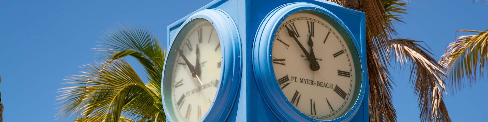 A blue clock with Roman numerals, labeled "Ft. Myers Beach," set against a clear blue sky with palm trees in the background.